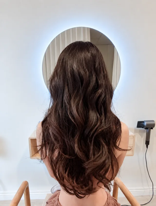 A client's long, glossy wavy hair after treatment, in front of the backlit round mirror.